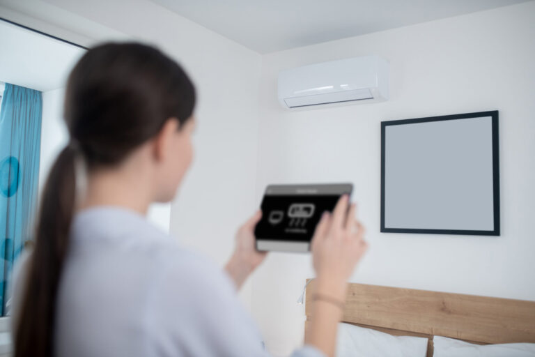 Back view of a dark-haired lady with the electronic device in the hands standing before the wall-mounted air conditioning unit